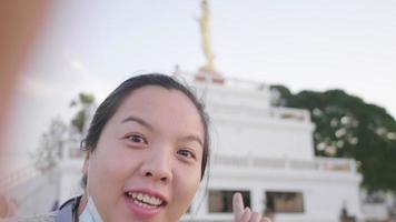 Close up of Asian woman standing and taking video selfie infront of monk statue at park. Looking at camera and talking about the view. monk statue background