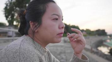 Nahaufnahme einer asiatischen Frau, die am Fluss sitzt und gutes Wetter bekommt, um etwas frische Luft zu bekommen. sitzen am river.trees hintergrund, schöne aussicht am abend. Wintersaison video