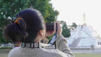 Back view of Asian woman standing and using camera to take a picture at park in beautiful day. Big tree and monk statue background. Relaxing day video