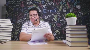 Front view of Asian man wearing glasses looking at camera to have a meeting online on laptop  and using wireless microphone on the table at home, serious talk. Trying to explain work. Working at home video