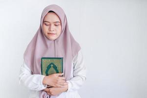 la joven islámica asiática que lleva un pañuelo en la cabeza sostiene el sagrado corán con los ojos cerrados y la cara tranquila. mujer indonesia sobre fondo gris foto