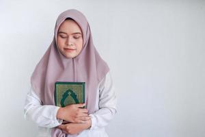 Young Asian Islam woman wearing headscarf is holding holy al quran with close eyes and calm face. Indonesian woman on gray background photo