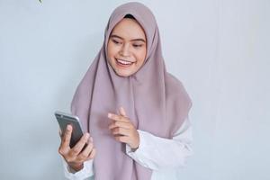 la joven islámica asiática que lleva pañuelo en la cabeza sonríe y es feliz con lo que ve en el smartphone. mujer indonesia sobre fondo gris foto