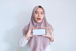 una joven islámica asiática con pañuelo en la cabeza se muestra en la pantalla blanca del teléfono celular en una vista horizontal con conmoción y sentimiento feliz. mujer indonesia sobre fondo gris foto