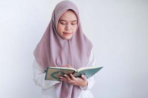 una joven islámica asiática que lleva un pañuelo en la cabeza reza o lee el corán, el libro sagrado del islam, con una sonrisa y una cara seria. mujer indonesia sobre fondo gris foto