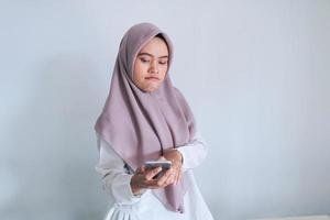 la joven islámica asiática que usa pañuelo en la cabeza está sorprendida, disgustada y enojada por lo que ve en el teléfono inteligente. mujer indonesia sobre fondo gris foto