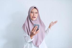 la joven islámica asiática que usa pañuelo en la cabeza está sorprendida, disgustada y enojada por lo que ve en el teléfono inteligente. mujer indonesia sobre fondo gris foto