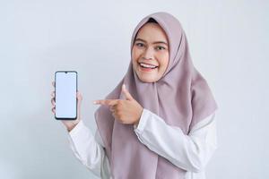 Young Asian Islam woman wearing headscarf is pointing finger on white screen of phone with smile and happy feeling. Indonesian woman on gray background photo