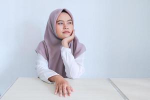 Young Asian Islam woman wearing headscarf is looking outside or daydreaming by holding the chin. Successful Islam Indonesian woman on gray background photo