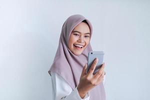 la joven islámica asiática que usa pañuelo en la cabeza está feliz y emocionada celebrando lo que ve en el teléfono inteligente. mujer indonesia sobre fondo gris