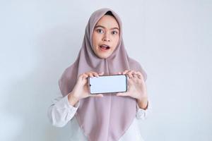 Young Asian Islam woman wearing headscarf is showing on white screen of cell phone in horizontal view with shock and happy feeling. Indonesian woman on gray background photo