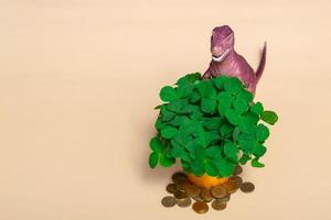 St. Patrick's day happy dinosaur, a bouquet of clover in a bucket and gold coins on a beige background. Closeup copy space photo