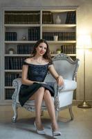 A beautiful woman with makeup and red lipstick is sitting in a black dress on a white chair against the background of shelves with books photo