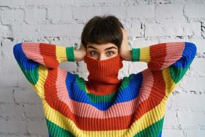 A young woman in a bright multicolored rainbow sweater hides her face and covers her ears with her hands. photo