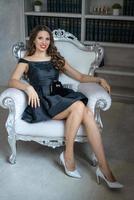 A beautiful woman with makeup and red lipstick is sitting in a black dress on a white chair against the background of shelves with books photo