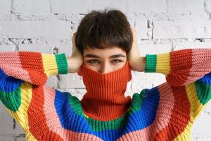 A young woman in a bright multicolored rainbow sweater hides her face and covers her ears with her hands. photo