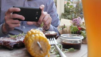 man neemt foto van eten op tafel - rijp varkensvlees, maïs, salade, bier - straatvoedsel video