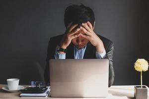 Stressed asian Businessman holding his head while looking for ideas with laptop, coffee cup, calculator on the book, decorative plants and book on wooden office desktop isolated on grey background photo