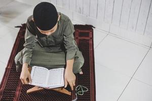 el retrato de un niño musulmán asiático vestido con un traje tradicional estaba leyendo el libro sagrado al-quran en la alfombra de oración. bandung, indonesia, diciembre de 2020 foto