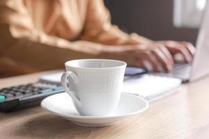 A cup of coffee on a desk office with blurry hand of businessman or worker typing on the laptop on the background