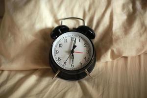 Top view of classic black alarm clock showing six o'clock with a bed in the background, waking up in the morning concept photo