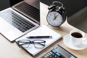 Business accessories on wooden office desk, laptop, glasses, pen, book, a cup of coffee, calculator and alarm clock photo