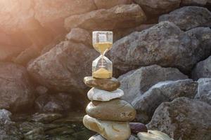 An hourglass with golden sand on sea rocks against a blue sea background. photo