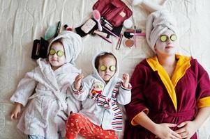 madre con hijas en un albornoz con una toalla en la cabeza cuida la piel en casa, pepinos en los ojos. foto