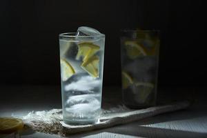 dos vasos de limonada helada con rodajas de limón y hielo en una servilleta de lino sobre fondo negro desdibujado. foto