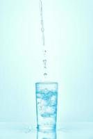 Pouring water into glass with ice isolated on blue background. Water spilled on the table. photo