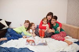 Happy big family is having fun together in bedroom. Large family morning concept. Mother with four kids wear pajamas read book in bed at home. photo
