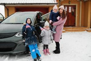 Young american mother with kids hold USA flags and charging electric car in the yard of her house at winter. photo