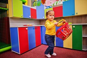 Cute baby girl playing in indoor play center. Kindergarten or preschool play room. Holds a basket for groceries in the children's kitchen.