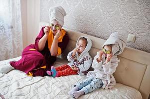 madre con hijas en un albornoz con una toalla en la cabeza cuida la piel en casa, pepinos en los ojos. foto