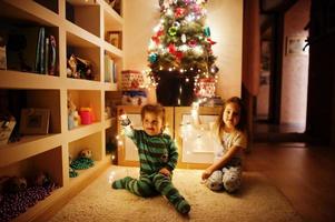 Two baby girl sisters looking on Christmas tree with shining garlands on evening home. photo