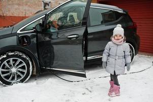 Baby girl against charging electric car in the yard of house at winter. photo