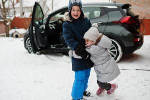 Brother with sister play against charging electric car in the yard of house at winter. photo