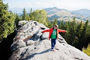 Boy on top of mountain. Children hiking on beautiful day in mountains, resting on rock and admire amazing view peaks. Active family vacation leisure with kids.Outdoor fun and healthy activity. photo