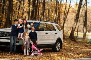 Mother stand near white suv car with four kids in autumn park. Family walk in fall forest. photo