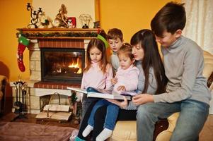 Happy young large family by a fireplace in warm living room on winter day. Mother with four kids at home read book. photo