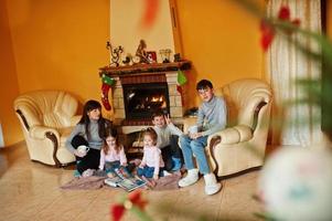 Happy young large family by a fireplace in warm living room on winter day. Mother with four kids at home read book.