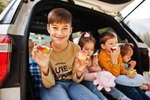 Family of four kids eat apples at vehicle interior. Children sitting in trunk. Traveling by car in the mountains, atmosphere concept.
