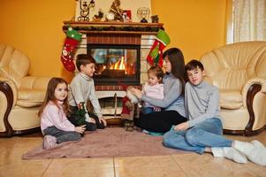 Happy young large family by a fireplace in warm living room on winter day. Mother with four kids at home.