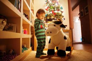 Baby girl on rocking cow toy against Christmas tree with shining garlands on evening home. photo