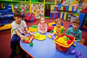 cuatro niños jugando en el centro de juegos interior. sala de juegos de jardín de infantes o preescolar. sentado junto a la mesa con frutas de plástico. foto