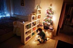 Baby girl on rocking cow toy against Christmas tree with shining garlands on evening home. photo