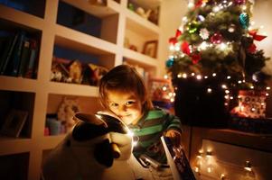 Baby girl on rocking cow toy against Christmas tree with shining garlands on evening home. photo