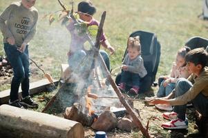 Family bonfire in mountain. Frying sausages. Mother with four kids camping. Autumn hike and camp weather. Warming and cooking near flame together. photo