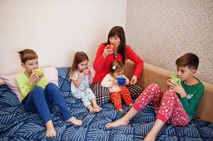 la gran familia feliz se está divirtiendo juntos en el dormitorio. gran concepto de mañana familiar. madre con cuatro hijos usa pijama bebe té en la cama en casa. foto