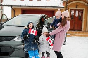 Young canadian mother with kids hold Canada flags and charging electric car in the yard of her house at winter. photo